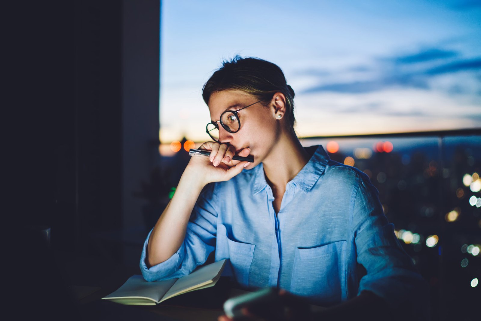Young concentrated female in casual clothes looking away sitting at table with notebook and pen touching face while considering idea for new business project at night